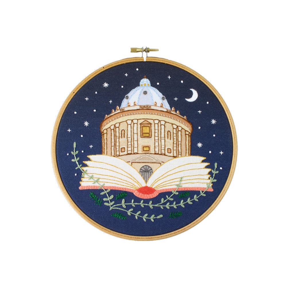 Embroidery hoop with a design of the Radcliffe Camera Library inside an open book against a night sky.