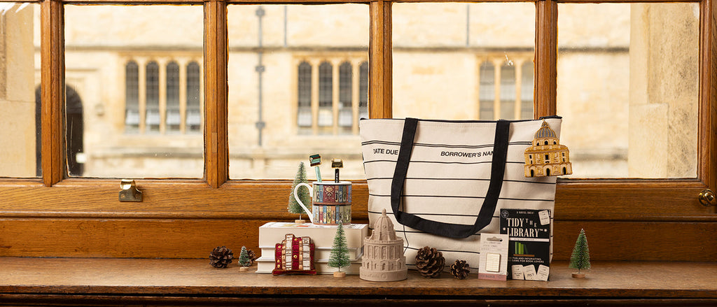 A collection of library themed items such as a library card bag, book covers design mug and book spines hanging decoration sit on a wooden window sill with view of the Bodleian Library visible through the window behind