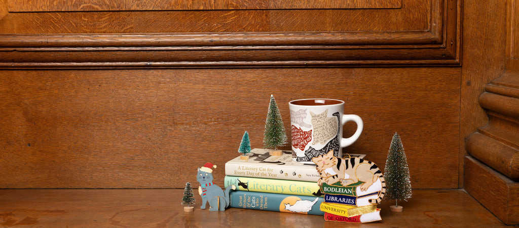 A collection of cat themed books, decorations and a mug are sat on a wooden shelf with a wood background