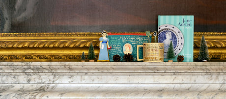 A Jane Austen decoration, pouch, mug, 2 pens and a book are sat on a marble mantlepiece with the bottom of a large gold frame visible in the background