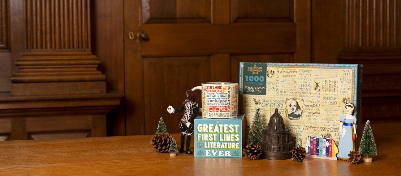 A collection of literary themed items including a mug, jigsaw, a model of the Radcliffe Camera and decorations of Jane Austen and Hamlet are sat on a wooden surface within a wood clad room