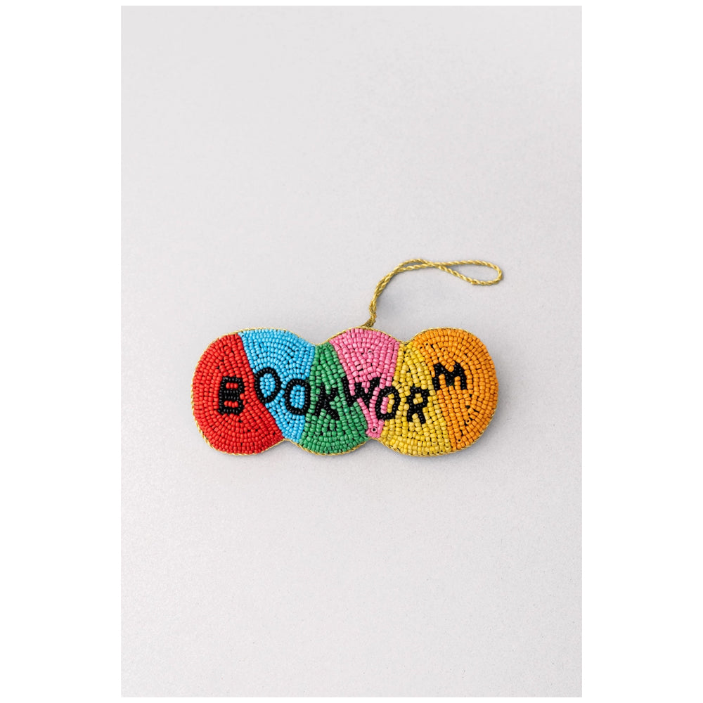 A colorful beaded ornament shaped like a worm with the word 'BOOKWORM' spelled out in black beads, hanging from a gold string on a light gray background.